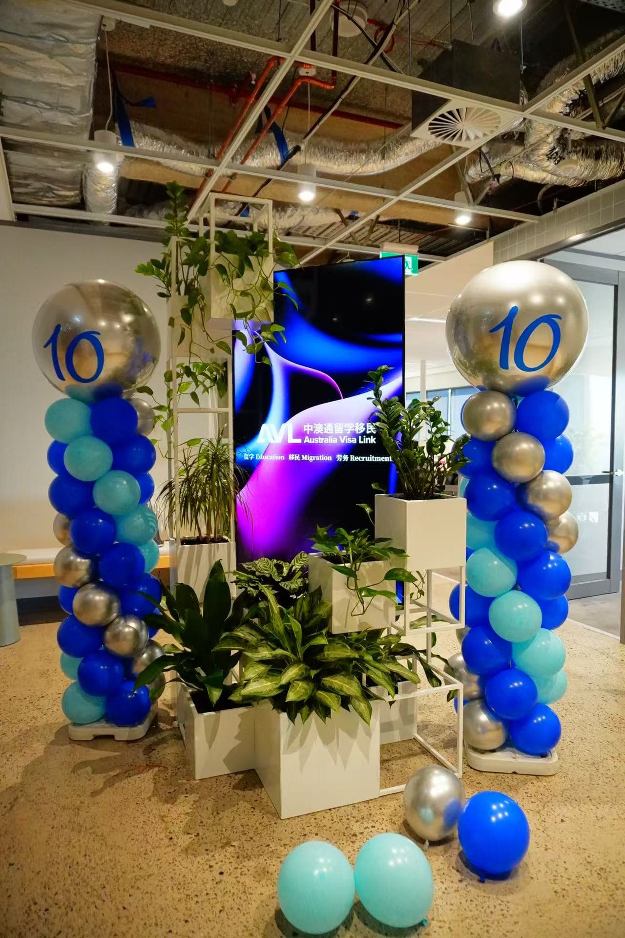Corporate 10th anniversary celebration featuring elegant blue and silver balloon columns with large "10" number balloons. Modern office space decorated with blue, teal, and silver color scheme, complemented by indoor plants and digital display.