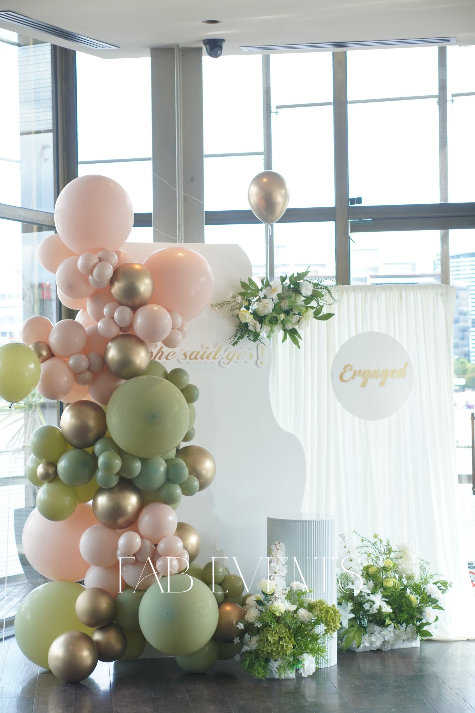 Romantic engagement setup with blush pink, sage green and gold balloon garland, white backdrops and floral arrangements