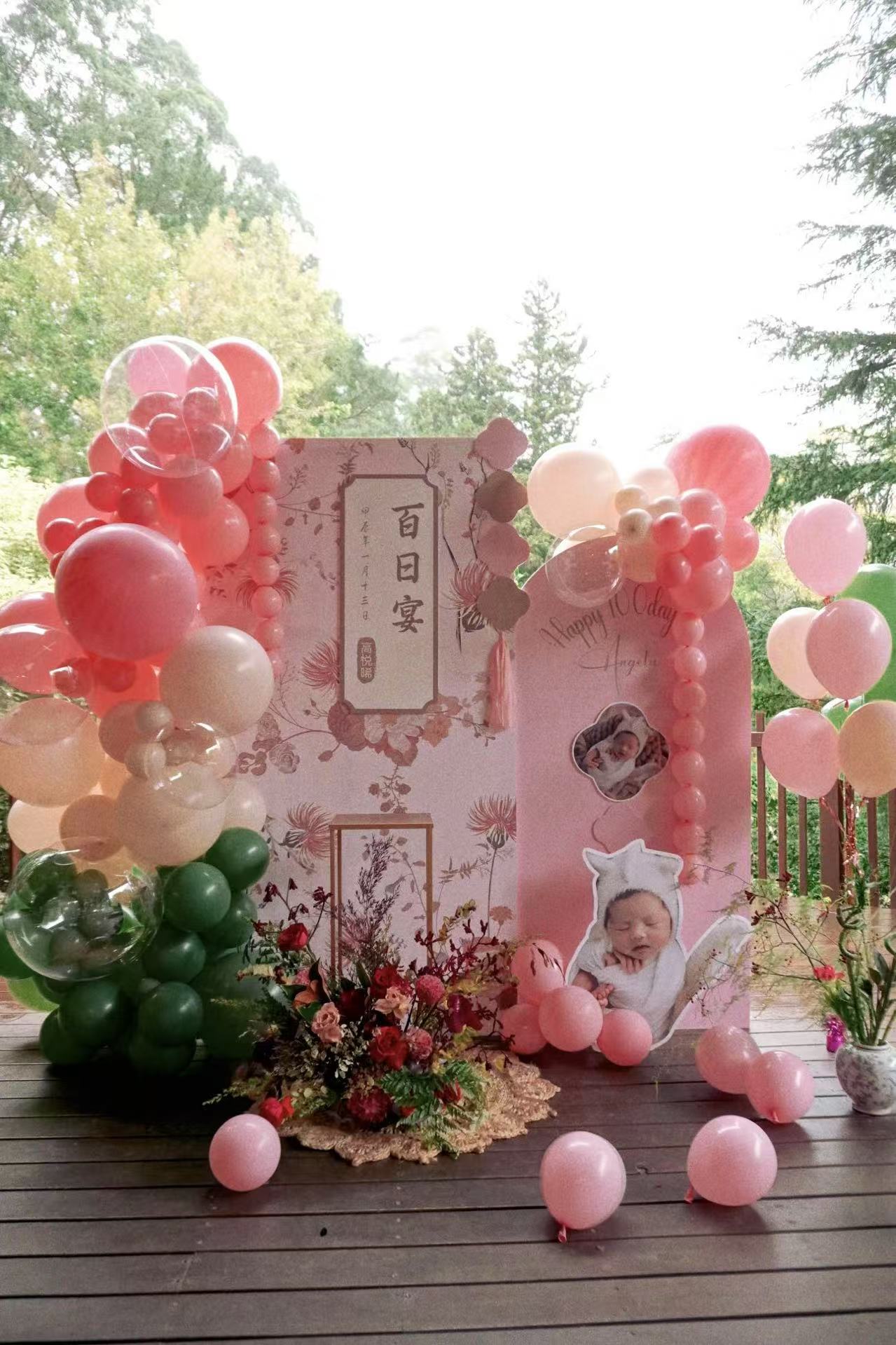 Stunning outdoor 100 days celebration featuring a pink floral themed backdrop with Chinese characters and baby photo display. Elaborate pink, white and green balloon arrangements frame the setup, complemented by fresh flower decorations at the base. Set on a wooden deck with natural greenery backdrop creating a perfect garden celebration atmosphere.