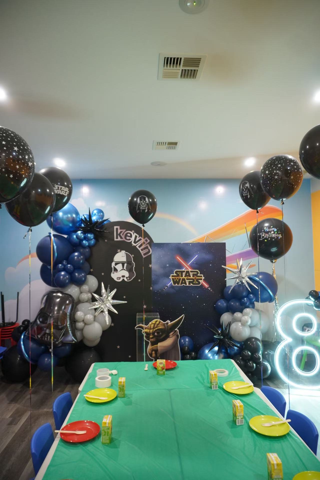 Epic Star Wars themed 8th birthday celebration for Kevin featuring a galaxy backdrop with Yoda character and Star Wars logo. Striking blue and black balloon arrangements with silver star accents, complemented by a neon number "8" light. Table setup includes green tablecloth with colorful plates for a complete themed experience.