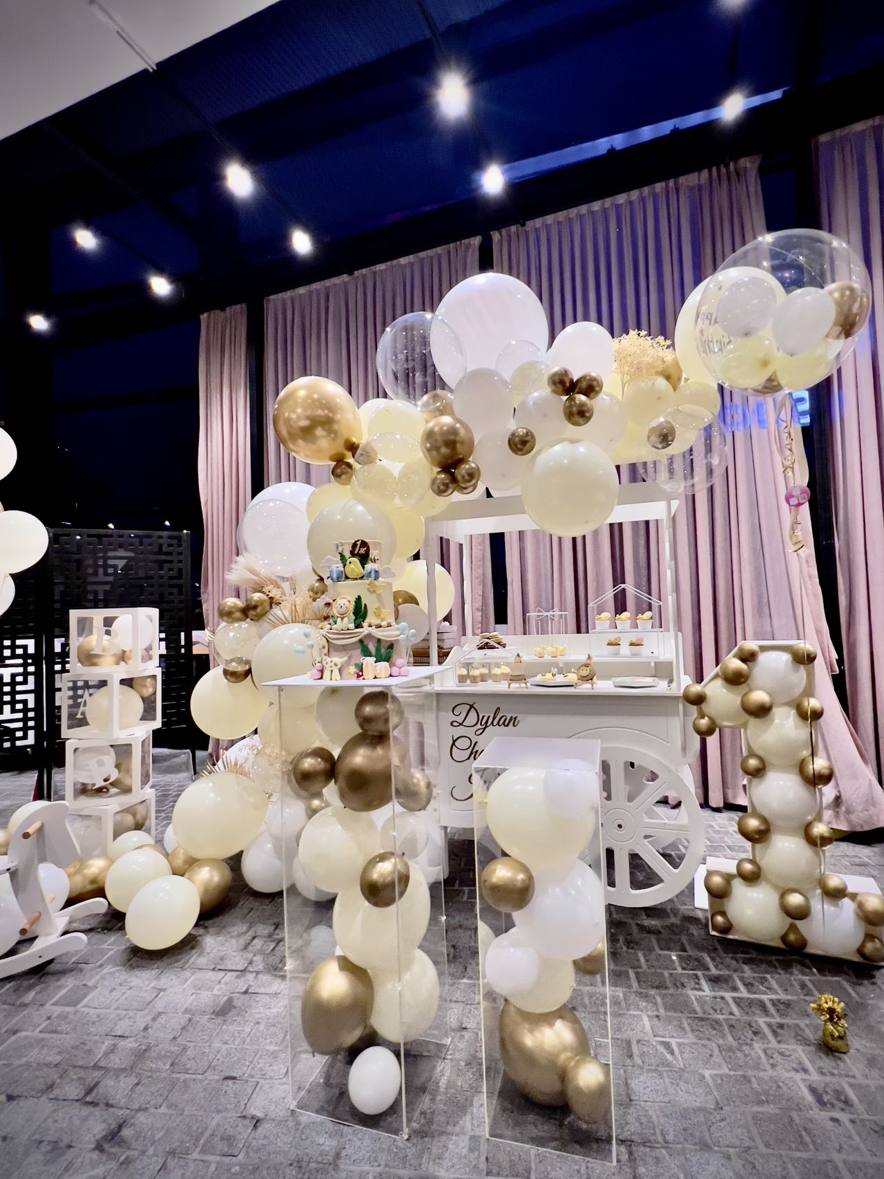 Elegant first birthday setup with white and gold balloons and dessert cart